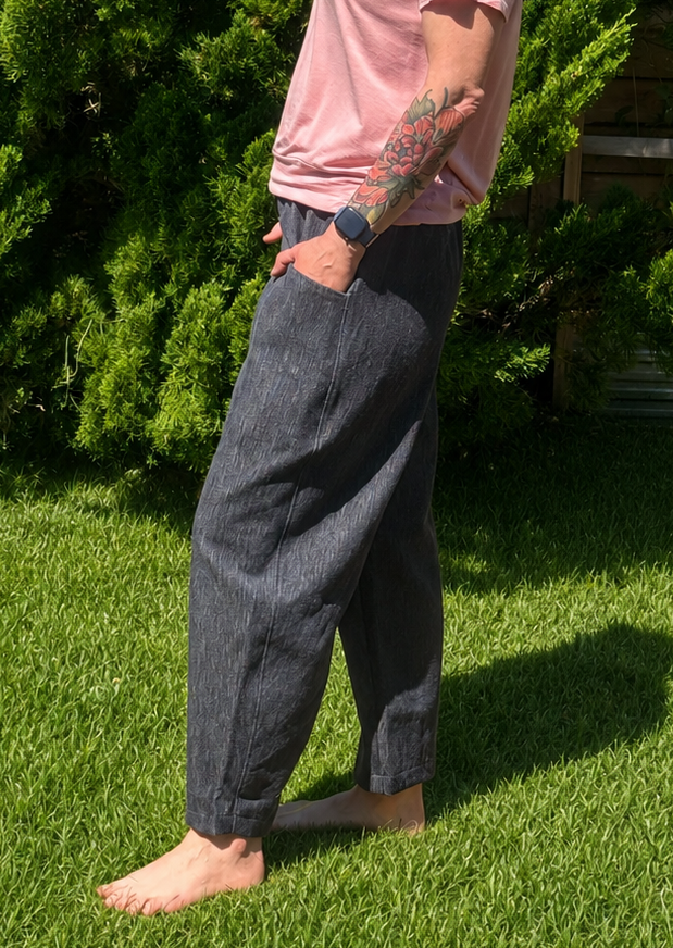 A person standing barefoot on green grass outdoors, wearing loose dark gray wide-leg pants and a pink t-shirt, with bushes in the background.