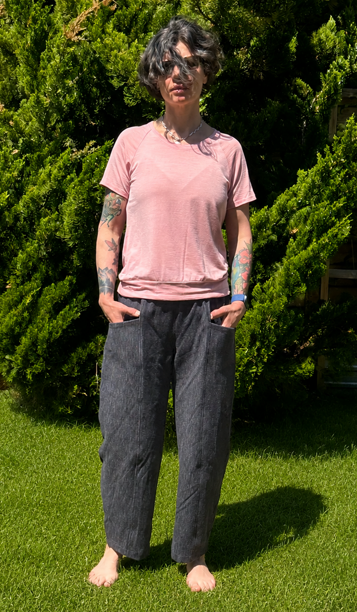 A person standing barefoot on a green lawn in front of tall green bushes, wearing a light pink short-sleeve t-shirt and dark charcoal gray wide-leg pants.