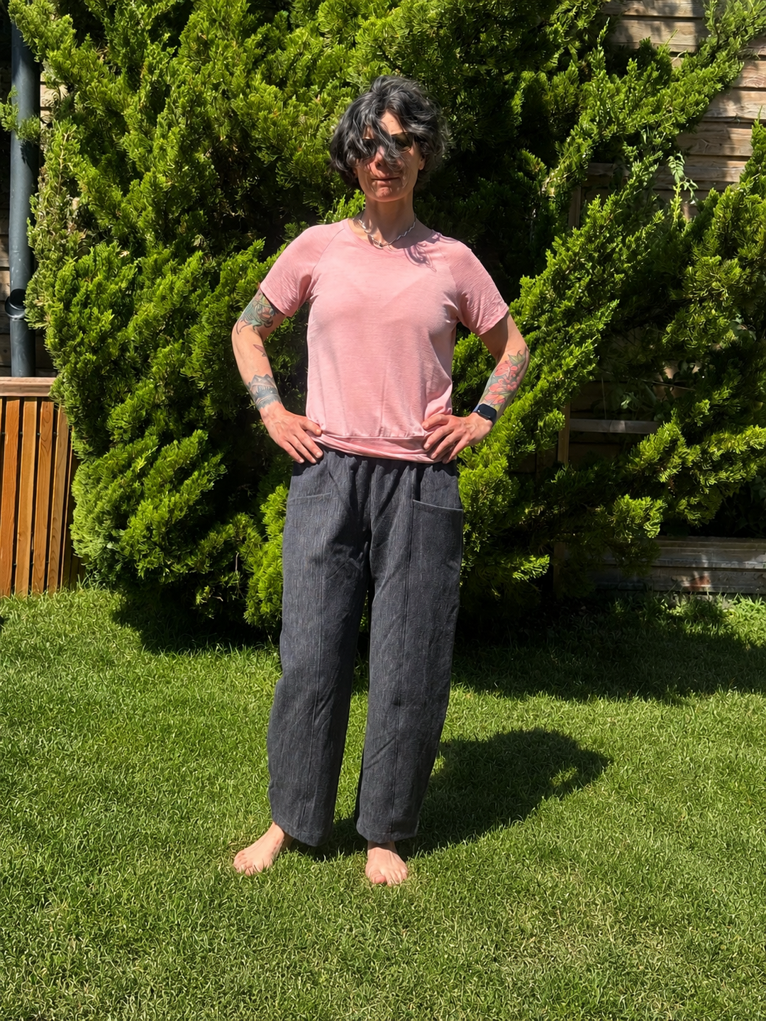 A person standing outdoors on a grassy lawn in front of green bushes, wearing a light pink short-sleeve t-shirt and dark charcoal gray wide-leg pants.