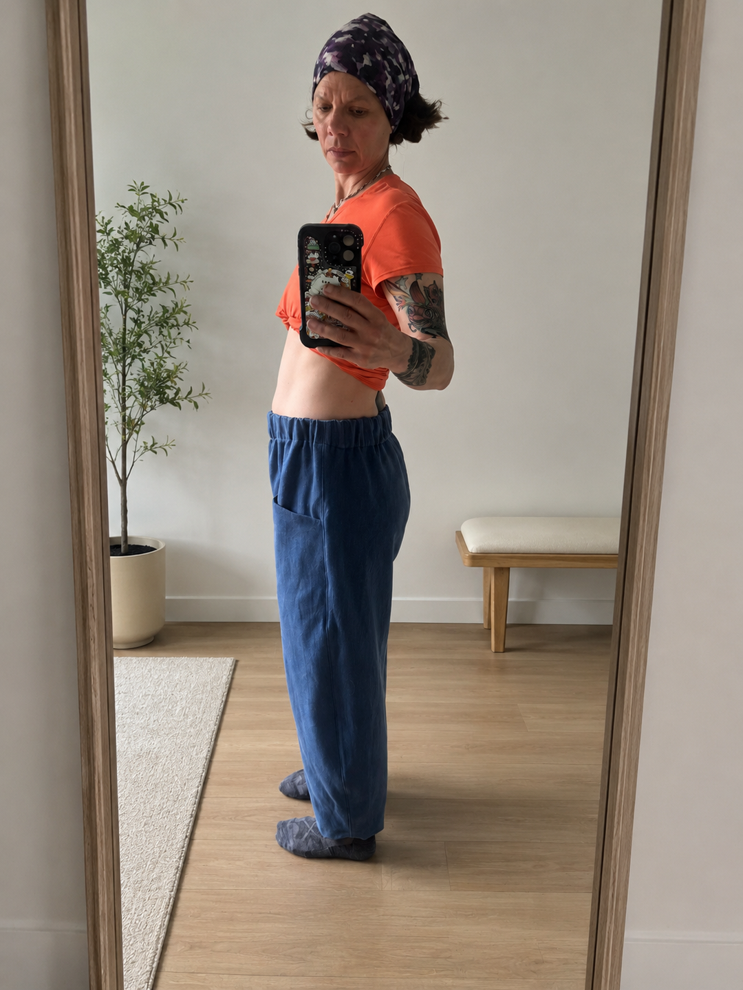 A person taking a mirror selfie in a room, wearing an orange short-sleeve crop top and loose blue pants. The room has light wood floors, a white wall, a potted plant, and a small bench.