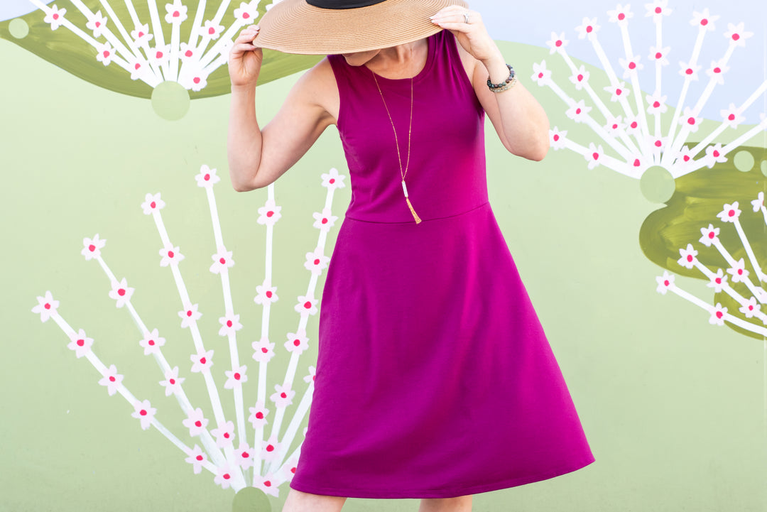 A woman wearing a purple fit and flare dress with short sleeves, styled with a wide-brimmed hat and a necklace. The background features a pattern of small white flowers.